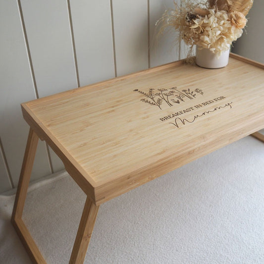 Wooden breakfast in bed tray with engraved text and floral design against a white wall.