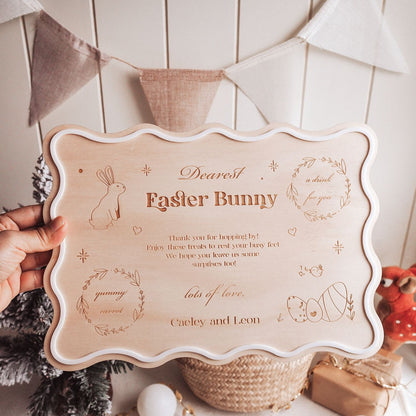 Wooden sign with engraved text and illustrations held by a hand, with Easter-themed decorations in the background.