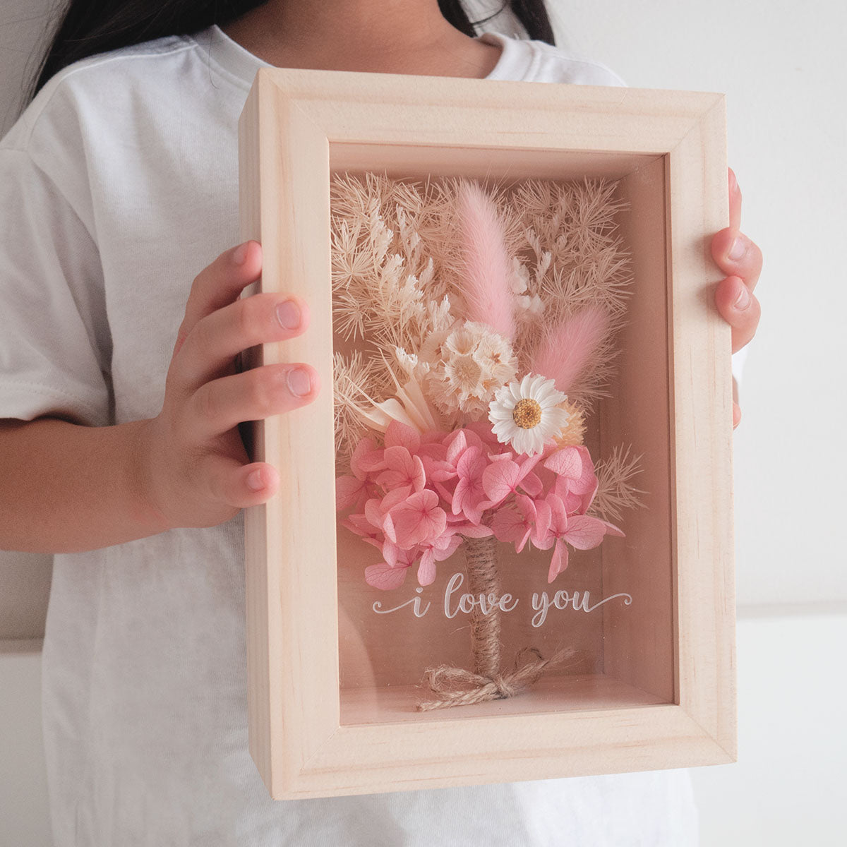 This is a wooden flower box with dried  pink  and white flowers and  grass. This is a front view with thext "i love you". A little girl is holding the box