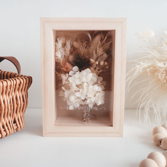 This is a wooden flower box with dried  natural brown and white flowers and  grass. This is a front view with thext "i love you"
