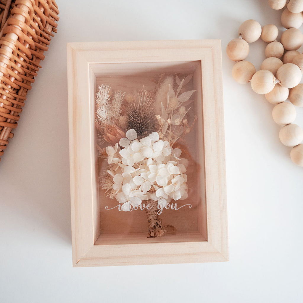 This is a wooden flower box with dried natural brown  and white flowers and  grass. This is a top view with the text "i love you".