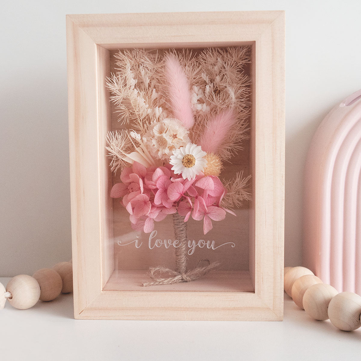 This is a wooden flower box with dried  pink  and white flowers and  grass. This is a front view wit thext "i love you"