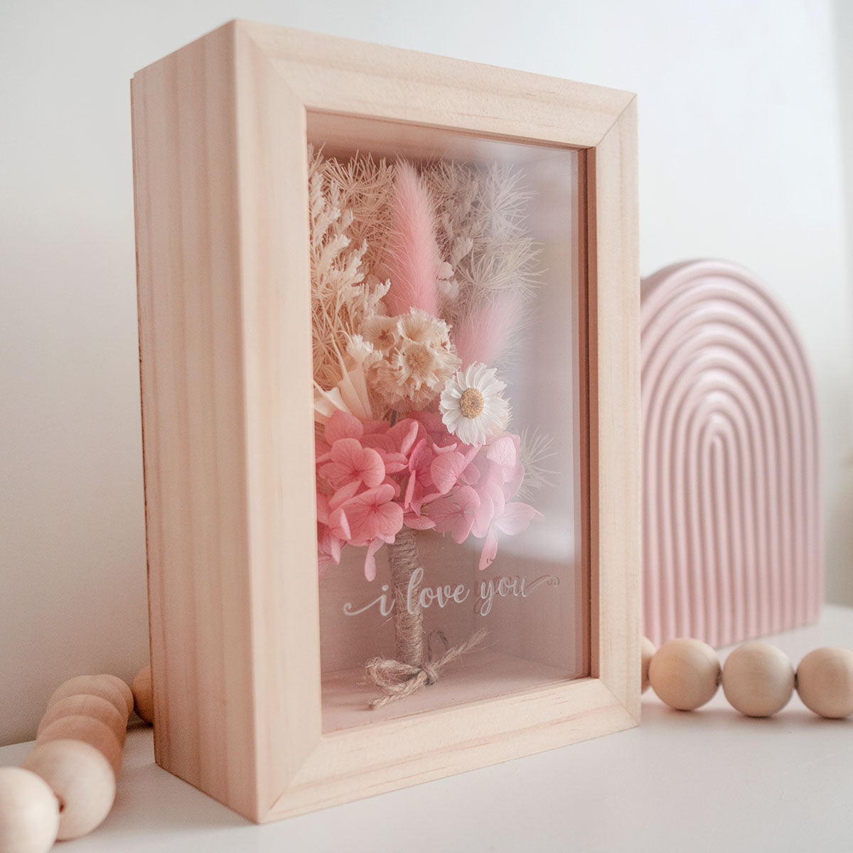 This is a wooden flower box with dried  pink  and white flowers and  grass. This is a side view with thext "i love you"