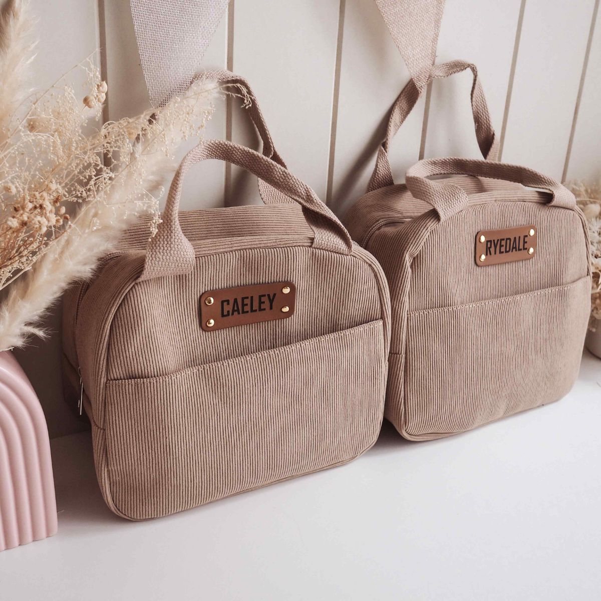 Two beige corduroy insulated lunch bags with personalized PU leather patches, one engraved with 'CAELEB' and the other with 'RYLDALE'.