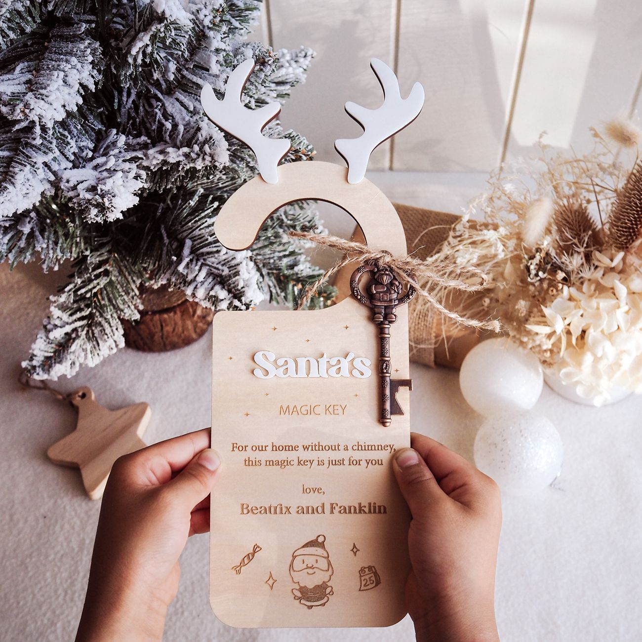 Wooden key-shaped ornament with reindeer antlers held by hands, Christmas tree and decorations in the background.