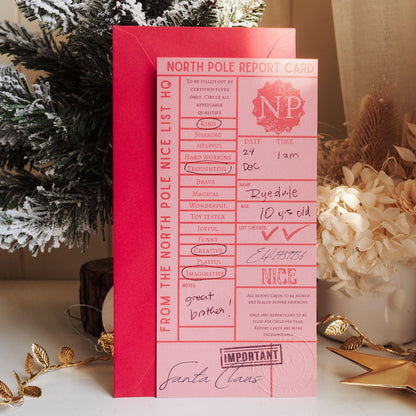 Pink 'North Pole Report Card' with Santa Claus signature on a decorative surface with Christmas elements.