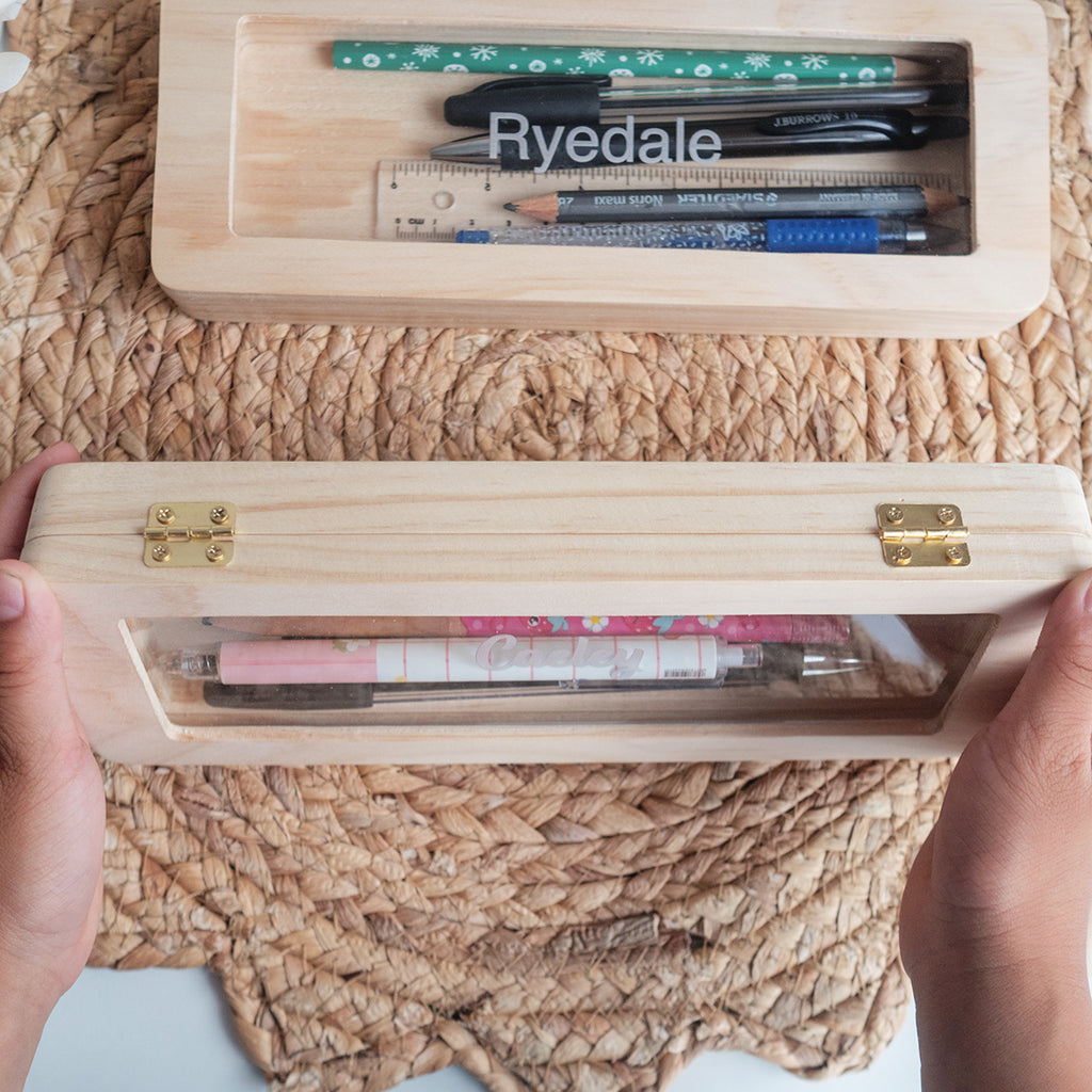 Personalised wooden pencil case made out of solid wood and clear acrylic. There's 2 pencil cases here with engraved names of Ryedale and Caeley. One of the pencil cases is showing the golden hinges
