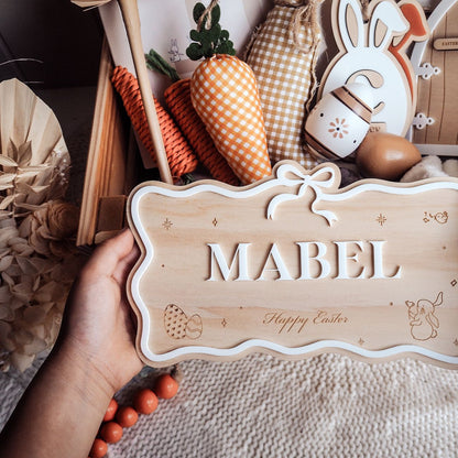 Hand holding a wooden sign with 'MABEL' and 'Happy Easter' text, surrounded by Easter decorations.
