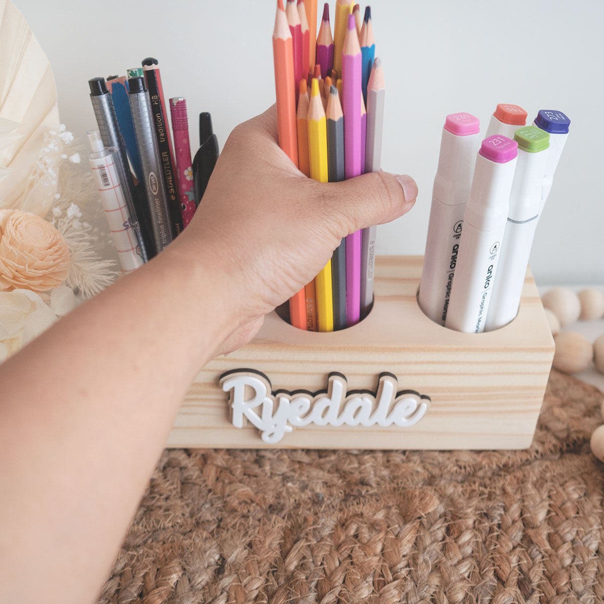 wooden pencil holder with three hole compartments. It is storing pencils, colour pencils and markers. It is also personalised. The photo is showing a hand grabbing the colour pencils