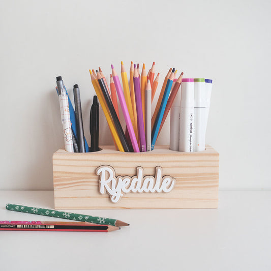 wooden pencil holder with three hole compartments. It is storing pencils, colour pencils and markers. It is also personalised. There's 2 pens on the table