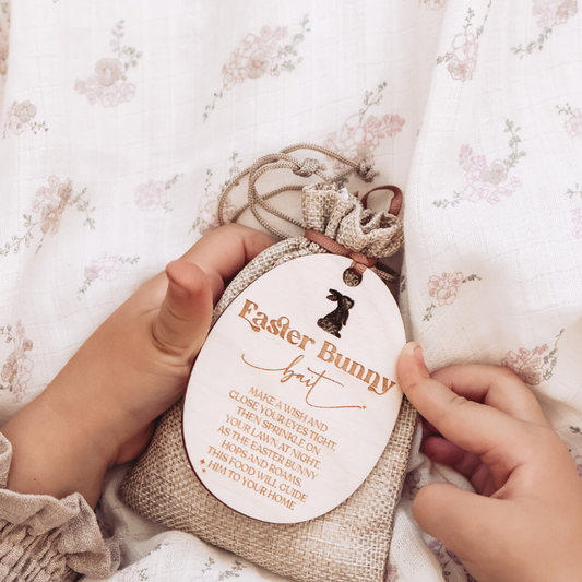 A child's hands holding a beige 'Easter Bunny Bait' bag with a tag attached, on a background with a floral pattern.