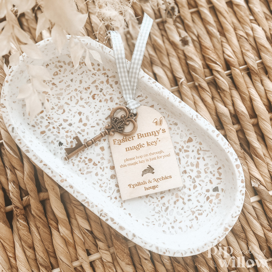 A wooden tag with a 'Easter Bunny Magic Key' and a metal key attached to it, displayed on a white plate, placed on a woven basket.