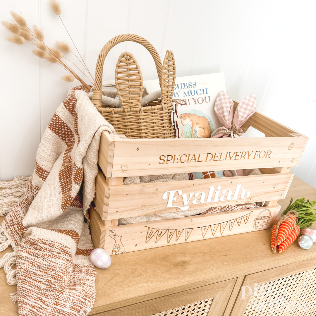 A wooden Easter crate with a handle, personalized with the name 'Tylalia', filled with Easter decorations and treats, placed on a table with a checkered cloth.