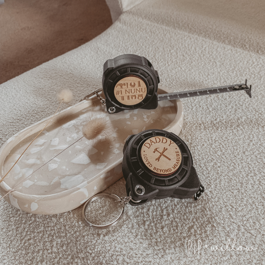 Personalised mini tape measures with keyrings, displayed on a white surface. One tape measure has 'DADDY' engraved on the front with a tool monogram design, and the other has a heart design.